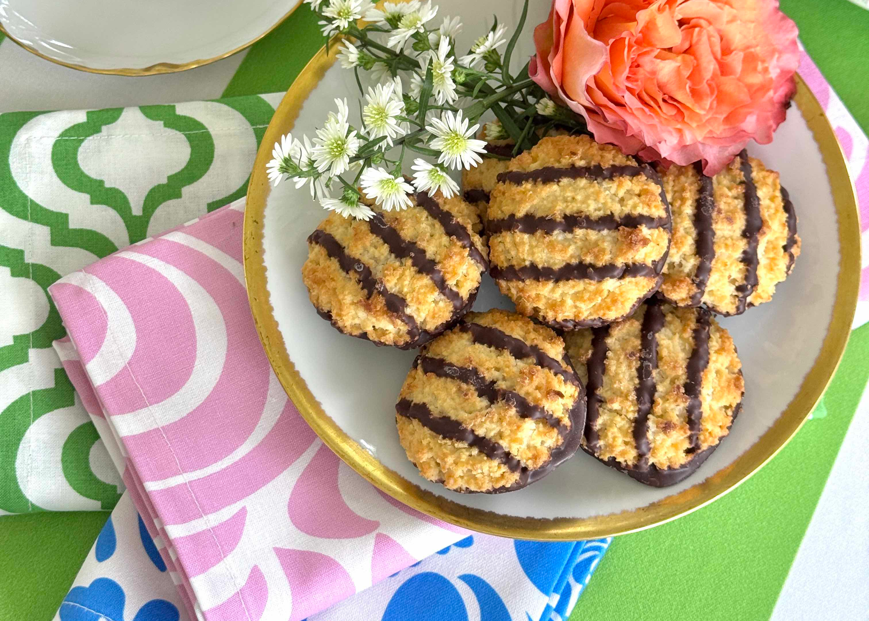 Five golden-brown coconut macaroon cookies with dark chocolate drizzle, arranged on a white ceramic plate with a gold rim. The plate is styled with white daisies and a pink rose, resting on a pink and white patterned napkin over a green and white patterned placemat.